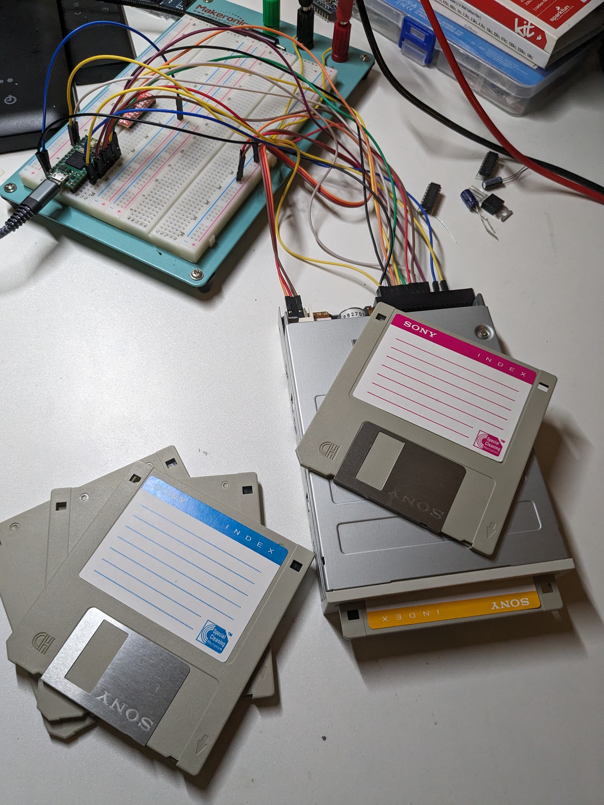 A blue floppy disk and a pink floppy disk laying around an ancient floppy drive that has yet a third floppy disk insertted into it. Behind them are various wires and circuitry which appears to be connected to the drive.